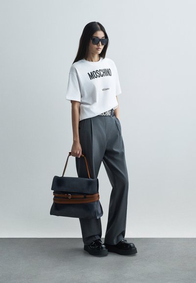 Kvinde iført hvid Moschino T-shirt, grå bukser, sorte plattform sko, solbriller, bærende en stor grå håndtaske med brune stropper.