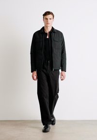 Schwarze gepolsterte Jacke mit zwei Brusttaschen, kombiniert mit schwarzen weiten Hosen und schwarzen Schuhen. Das Model steht vor einem schlichten Hintergrund.