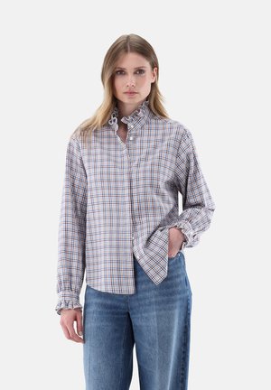 Woman wearing a high-neck ruffled plaid shirt with long sleeves and blue jeans, posing with one hand in pocket against a plain background.