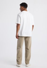 Joven con cabello rizado, vistiendo una camisa blanca de manga corta, pantalones beiges, zapatillas blancas y una cadena de oro, de pie de espaldas sobre un fondo blanco.