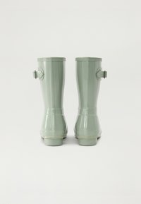 Bottes de pluie en caoutchouc vert avec une finition brillante, hauteur mi-mollet, et détail de sangle ajustable sur le côté. Texture lisse et forme arrondie.