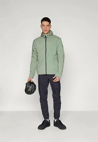 Light green, zippered jacket with hood, paired with dark pants and black shoes. Model holds a black bike helmet. Smooth, casual design.