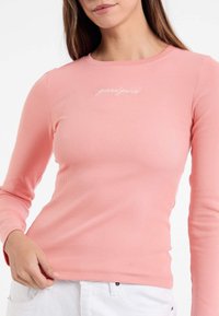 Šviesiai rožinė ilgarankovė tekstūruota palaidinė su apvalia iškirpte ir baltu išsiuvinėtu "guessjeans" logotipu krūtinėje. Minkšta tekstūra.