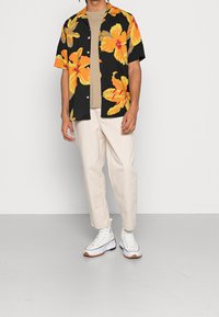 Zwart T-shirt met korte mouwen en een groot geel-oranje bloemenpatroon, gecombineerd met lichtbeige broek en witte hoge sneakers.