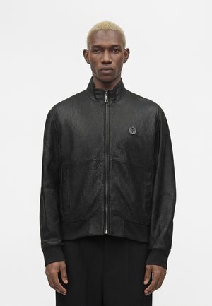 Man met kort blond haar draagt een zwarte getextureerde jas met rits en zwarte broek, staand tegen een effen witte achtergrond.