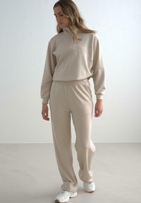 Beige Baumwollmischung Trainingsanzug mit einem Pullover mit halbem Reißverschluss, Hose mit elastischem Bund und lässiger Passform, kombiniert mit weißen Sportschuhen.