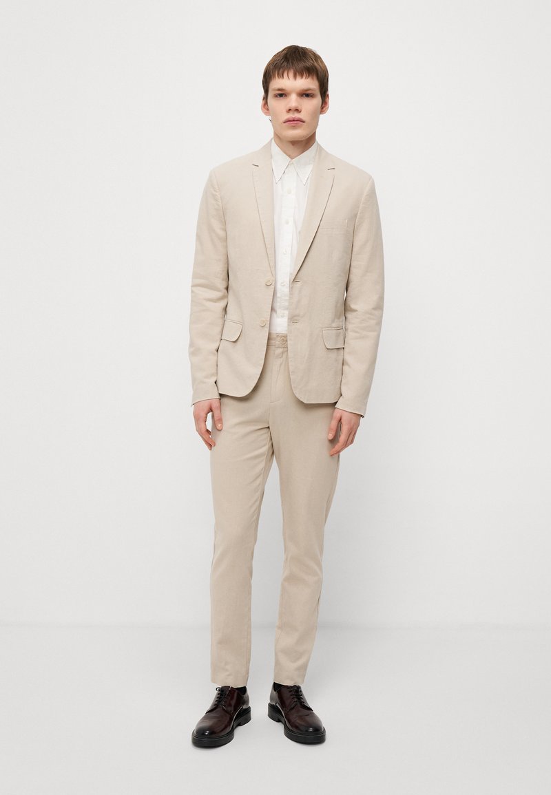 Costume beige ajusté en tissu léger, comprenant une veste à un bouton avec deux poches et un pantalon assorti, associé à une chemise blanche.
