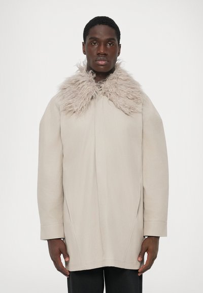 Jeune homme portant un manteau beige clair avec un large col duveteux, se tenant devant un fond blanc uni.