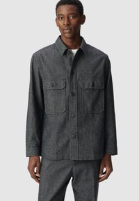 Giacca camicia tessuta grigio scuro, con collare a ribattini, tasche frontali e chiusura con bottoni. Maniche lunghe con polsini abbottonati.