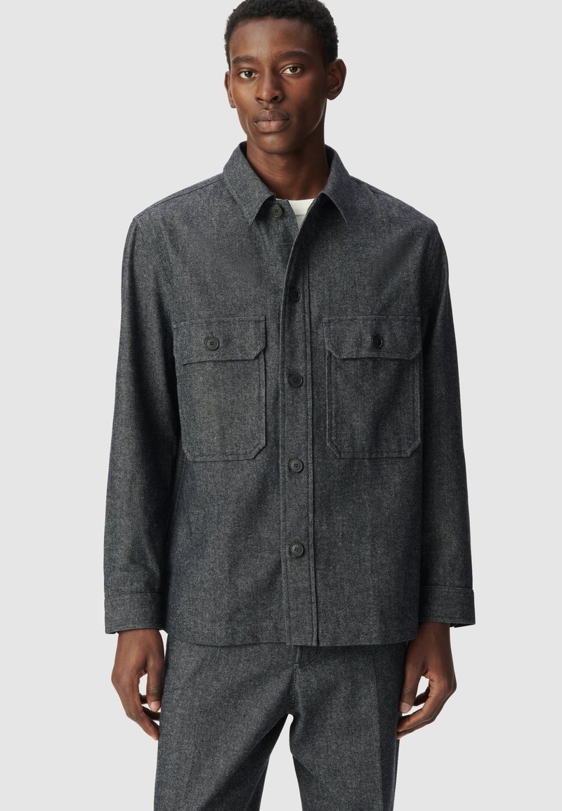 Giacca camicia tessuta grigio scuro, con collare a ribattini, tasche frontali e chiusura con bottoni. Maniche lunghe con polsini abbottonati.