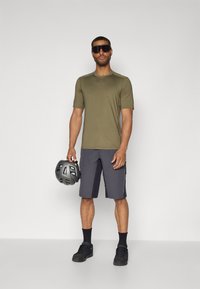 Chemise vert olive à manches courtes, shorts gris, baskets noires et lunettes de soleil. Tenant un casque gris avec des trous de ventilation.