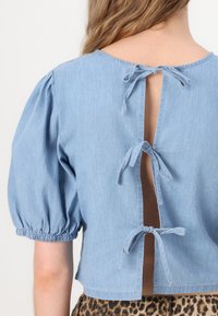 Ljusblå denimblus med puffärmar, öppen ryggdesign som säkras med tre knytningar, och en kort passform. Har en subtil textur.
