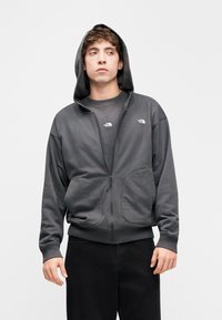 Γκρι φούτερ με φερμουάρ, κουκούλα και τσέπες καγκουρό. Μαλακό ύφασμα, άνετη γραμμή, με λογότυπα στο στήθος και στο μανίκι. Μαύρο παντελόνι.