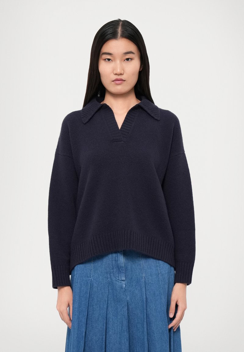 Navy knit sweater with a collar, v-neck design, and ribbed hem. Pair of blue pleated denim skirt visible below the sweater.