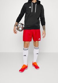 Sweatshirt à capuche noir, short rouge, chaussettes blanches avec des rayures noires, et chaussures de football orange. Ballon de football tenu dans une main.