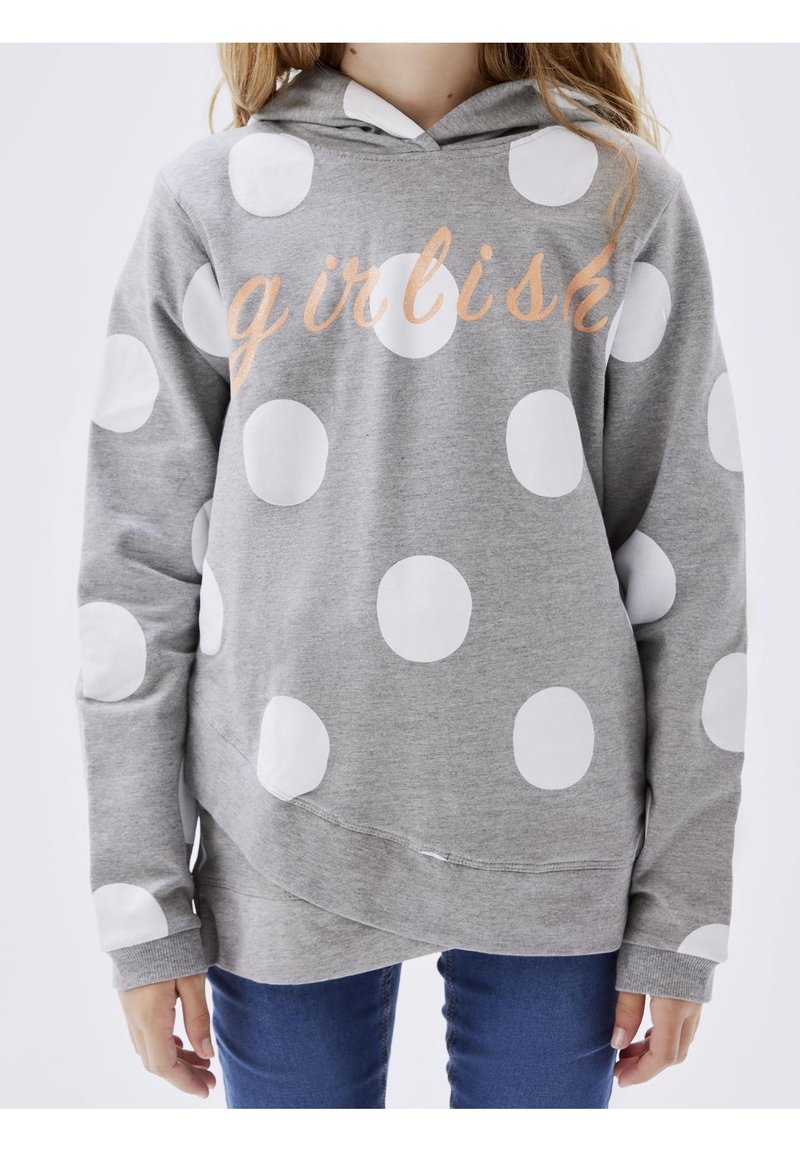 Enfant portant un sweat à capuche gris avec de gros pois blancs et le texte « girlish » en écriture cursive, associé à un jean bleu.