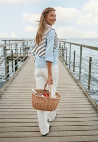 Femme souriante marchant sur un ponton en bois au-dessus de l'eau, vêtue de vêtements légers et portant un panier de pique-nique avec de la nourriture et des boissons.