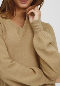 Pulover bej cu decolteu în V, fabricat dintr-un material moale, având mâneci puff și manșete ribbed, cu o croială lejeră. Țesătură texturată cu un luciu subtil.