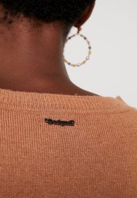Pull en tissu tricoté doux et chaud de couleur orange avec une texture côtelée. Il présente une étiquette logo noire au niveau de l'encolure arrière. Des boucles d'oreilles circulaires sont visibles.