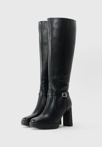 Botas negras de cuero hasta la rodilla con tacones altos y gruesos, y una correa decorativa con hebilla plateada alrededor del tobillo.