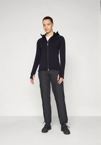 Nuori nainen seisoo valkoista taustaa vasten päällään musta vetoketjullinen huppari, mustat housut ja mustat vaelluskenkä, katsoen suoraan eteenpäin kädet rentoina.