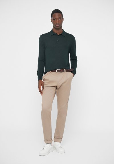 Σκούρο πράσινο πουκάμισο με μακριά μανίκια, μπεζ παντελόνι, πλεγμένη καφέ ζώνη και λευκά αθλητικά παπούτσια. Το μοντέλο στέκεται σε μια απλή λευκή φόντο.