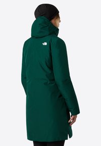 Grøn isoleret parka med hætte, der har et logo på ryggen, og en længere pasform. Glat tekstur og lige silhuet med sidelæg.