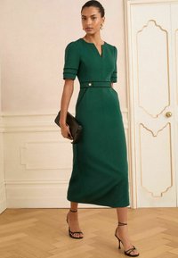 Femme portant une robe midi verte avec des manches bouffantes et des bordures noires, tenant une pochette noire, marchant dans une pièce aux teintes claires avec un sol en bois.