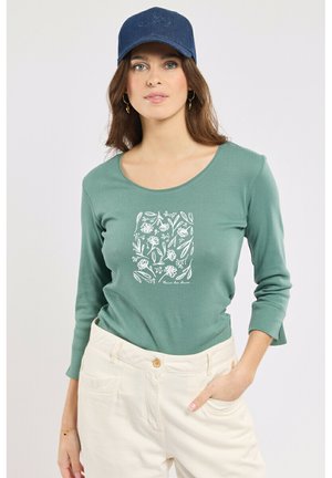 Femme portant un chemisier à manches longues couleur sarcelle avec un motif floral blanc, un pantalon crème, une casquette marine et des boucles d'oreilles créoles dorées, posant avec une main dans la poche.