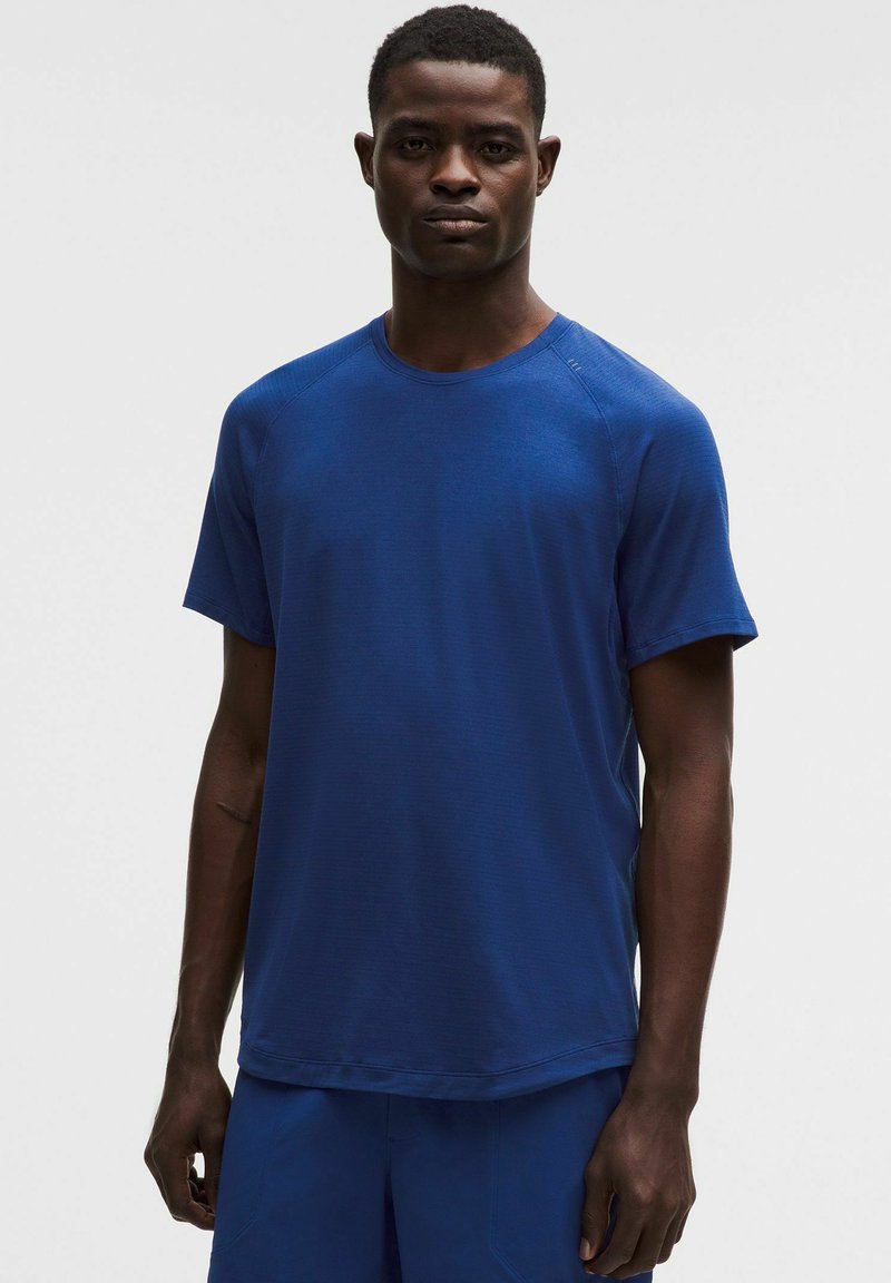 Mladić s kratkom kosom, odjeven u jednobojnu plavu kratku majicu s kratkim rukavima i odgovarajuće plave hlače, stoji na jednostavnoj svijetloj pozadini.