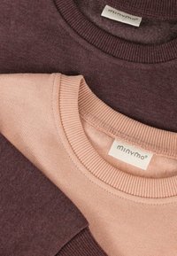 Dos sudaderas de cuello redondo dobladas y superpuestas, una de color rosa claro y otra de color burdeos oscuro, ambas con cuellos de canalé y etiquetas de la marca "minymo" visibles.