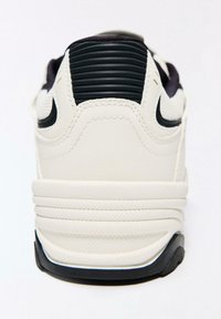 Fehér sportcipő texturált bőrfelsőrésszel, bordázott fekete díszítésekkel a saroknál, és gumitalppal, amely látható tapadási mintázattal rendelkezik.