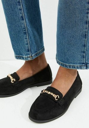 Mocassins en daim noir avec détail de chaîne dorée portés avec un jean en denim bleu, vus de dessus sur un sol blanc.
