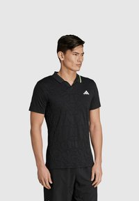 Zwarte poloshirt met een textuurontwerp en een groene accent op de kraag. Voorzien van het Adidas-logo op de linkerborst. Korte mouwen.