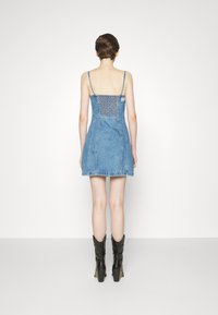 Robe en denim mini avec des bretelles spaghetti, un dos smocké et un ourlet effiloché. Associée à des bottes de cow-boy à talons noirs. Couleur bleu clair.
