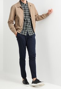 Vaaleanruskea bomber-takki vihreänruudullisen paidan päällä, laivastonsiniset slim-fit-housut ja mustat tennarit valkoisilla pohjilla. Asu on rento ja moderni.