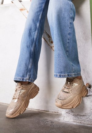 Nogi w jasnoniebieskich dżinsach noszące beżowe masywne sneakersy z ozdobami z kryształków, stopy unoszące się nad betonową podłogą i białą ścianą.