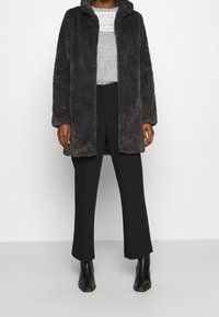 Šedý kabát z umělé kožešiny s texturovaným vlnitým vzorem, otevřeným předním dílem a velkým límcem, spárovaný se světle šedým topem a černými kalhotami s širokými nohavicemi.