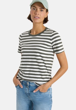 Femme portant un t-shirt à manches courtes rayé blanc et gris foncé, un jean bleu clair et une casquette de baseball vert clair, les mains dans les poches.