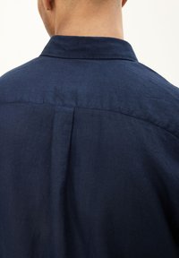 Camicia in lino blu navy con colletto strutturato, dettaglio della piega posteriore e una consistenza liscia, che mette in risalto il tessuto traspirante dell'indumento.