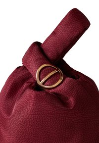 Burgundy fabric bag with a textured surface, featuring a gold circular metal accent at the top and a folded strap design.