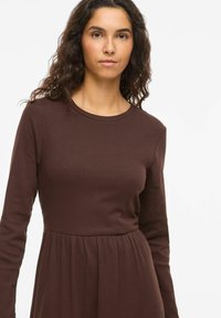 Robe marron à manches longues côtelées avec un col rond, un corsage ajusté et une jupe fluide plissée à la taille. Texture lisse.