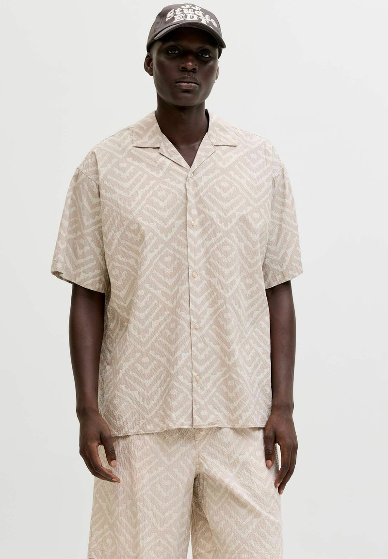 Personne à la peau foncée portant une chemise beige à manches courtes à motifs, un short assorti et une casquette de baseball foncée avec un texte blanc.