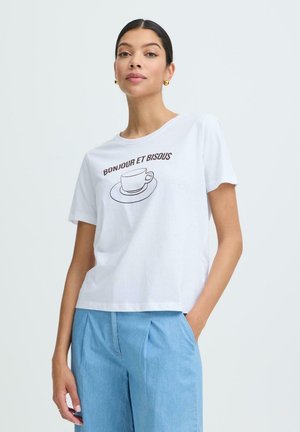 Femme aux cheveux bruns attachés, portant des boucles d'oreilles en or, un t-shirt blanc avec l'inscription "Bonjour et Bisous" et un motif de tasse de café, ainsi qu'un pantalon bleu clair.