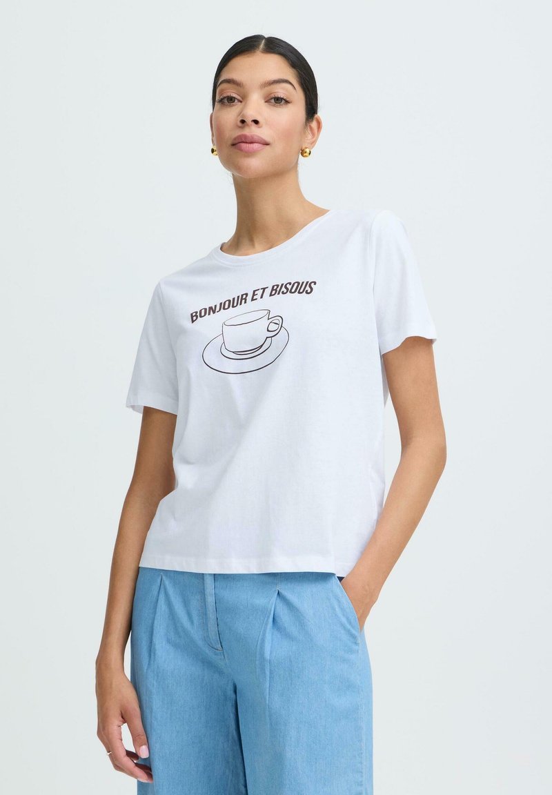 Femme aux cheveux bruns attachés, portant des boucles d'oreilles en or, un t-shirt blanc avec l'inscription "Bonjour et Bisous" et un motif de tasse de café, ainsi qu'un pantalon bleu clair.
