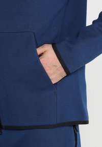 Veste en polaire bleue avec une poche. Présente des bordures noires le long des bords, une texture lisse et un léger éclat sur le tissu.
