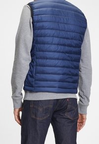 Marineblaue Steppweste mit horizontalen Nähten, über einem grauen Sweatshirt getragen. Seitennähte sind sichtbar. Dunkle Jeans runden das Outfit ab.