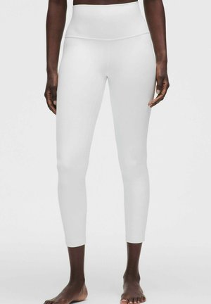 Person iført højtaljede, korte hvide leggings stående barfodet mod en ensfarvet lys baggrund.