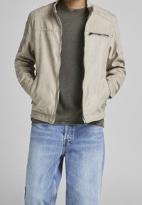 Beige zipová bunda s ribbovaným límcem, texturovanými rameny a náprsní kapsou, nošená přes tmavě zelenou košili a světle modré džíny.