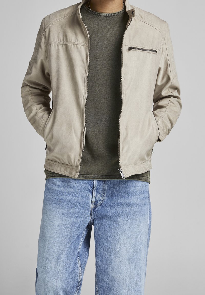 Beige zipová bunda s ribbovaným límcem, texturovanými rameny a náprsní kapsou, nošená přes tmavě zelenou košili a světle modré džíny.
